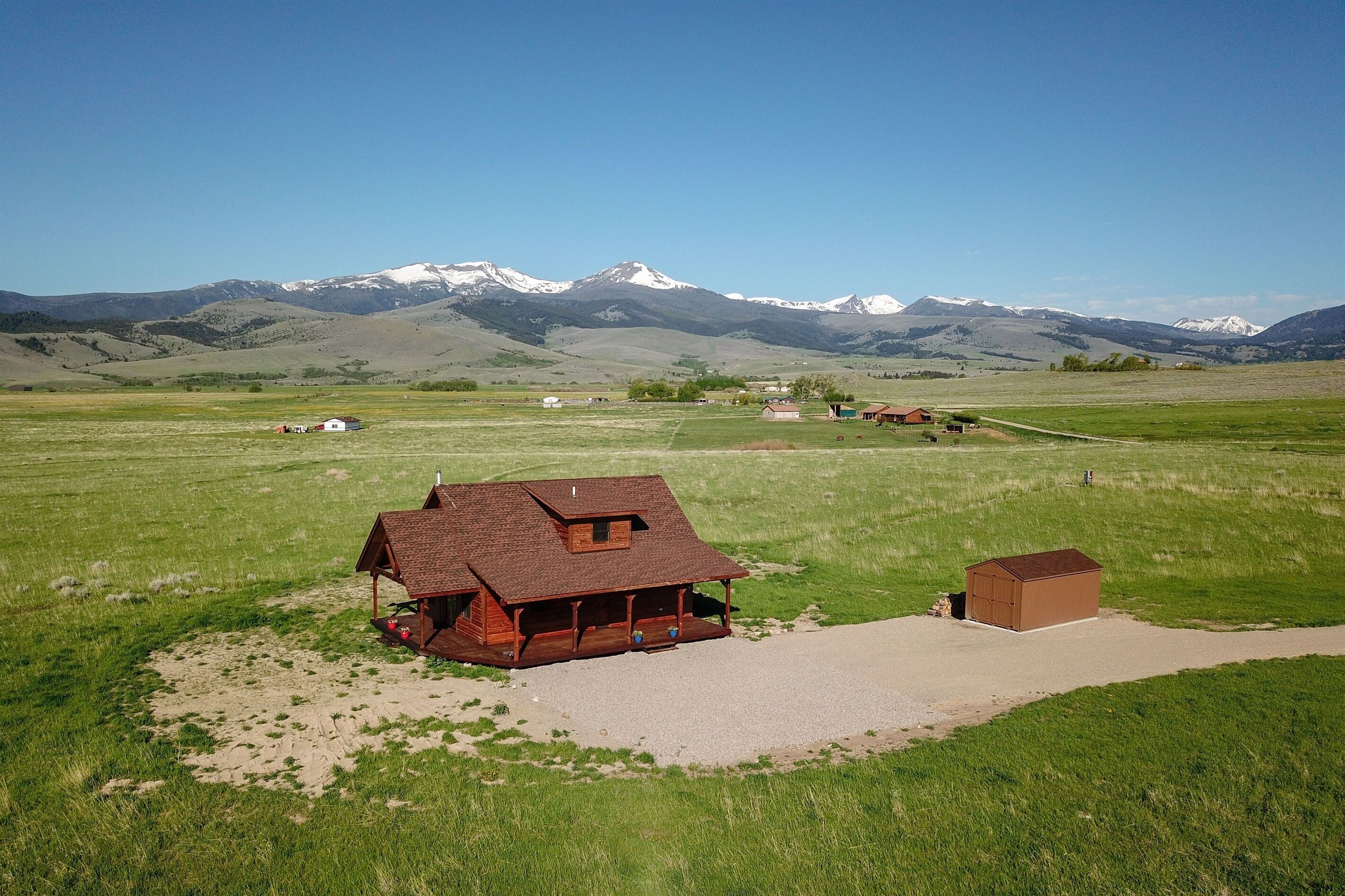 A wooden cabin with a porch, surrounded by a grassy field and a mountain range in the background.