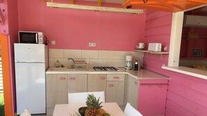 Eine Küche mit rosafarbenen Wänden, einem weißen Kühlschrank, einer Mikrowelle, einem Gasherd und einer Kaffeemaschine. In der Mitte steht ein Tisch mit einer Obstschale.
