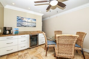 A kitchen with a coffee maker, a refrigerator, and a dining area with wicker chairs and a glass table.