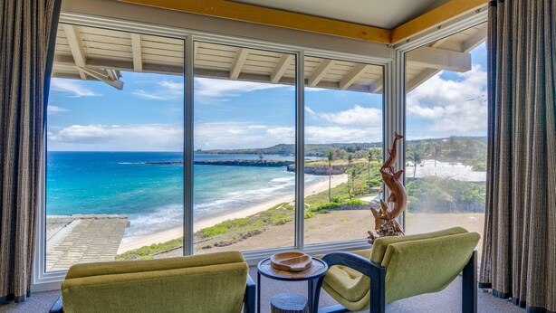 Habitación con una ventana grande que da a una playa y al océano, con dos sillas verdes y una pequeña mesa de madera.