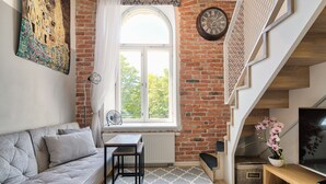 A cozy living room with a grey sofa, a small table, a window with a view, a brick wall, and a wooden staircase.