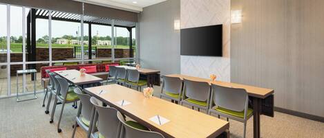 A conference room with tables and chairs, a large window with a view of greenery, and a flat-screen TV mounted on the wall.