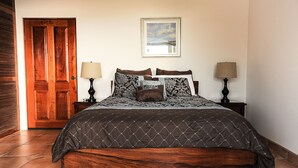 A bedroom with a wooden bed, two bedside lamps, a framed picture on the wall, and a wooden door. - Ocean View Single Story House with Private Infinity Pool.  10 min walk to Beach (Playas del Coco)