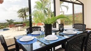 A dining area with a table set for four, large windows offering a view of palm trees and a pool. - Ocean View Single Story House with Private Infinity Pool.  10 min walk to Beach (Playas del Coco)