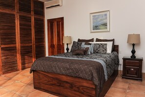 Een slaapkamer met een houten bed, twee nachtkastjes met lampen, een houten garderobe, een schilderij aan de muur en een betegelde vloer.