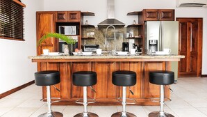 A kitchen with wooden cabinets, a central island with stools, a microwave, a coffee maker, and a refrigerator. - Ocean View Single Story House with Private Infinity Pool.  10 min walk to Beach (Playas del Coco)