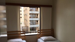 A room with two single beds, a window with a view of a building, and a wooden headboard. - Cozy, well-furnished apartment on a quiet street near the beach. (Guaruja)