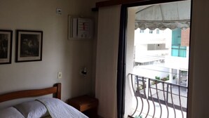 A bedroom with a bed, a nightstand, a wall-mounted thermostat, and a window with a view of buildings. - Cozy, well-furnished apartment on a quiet street near the beach. (Guaruja)