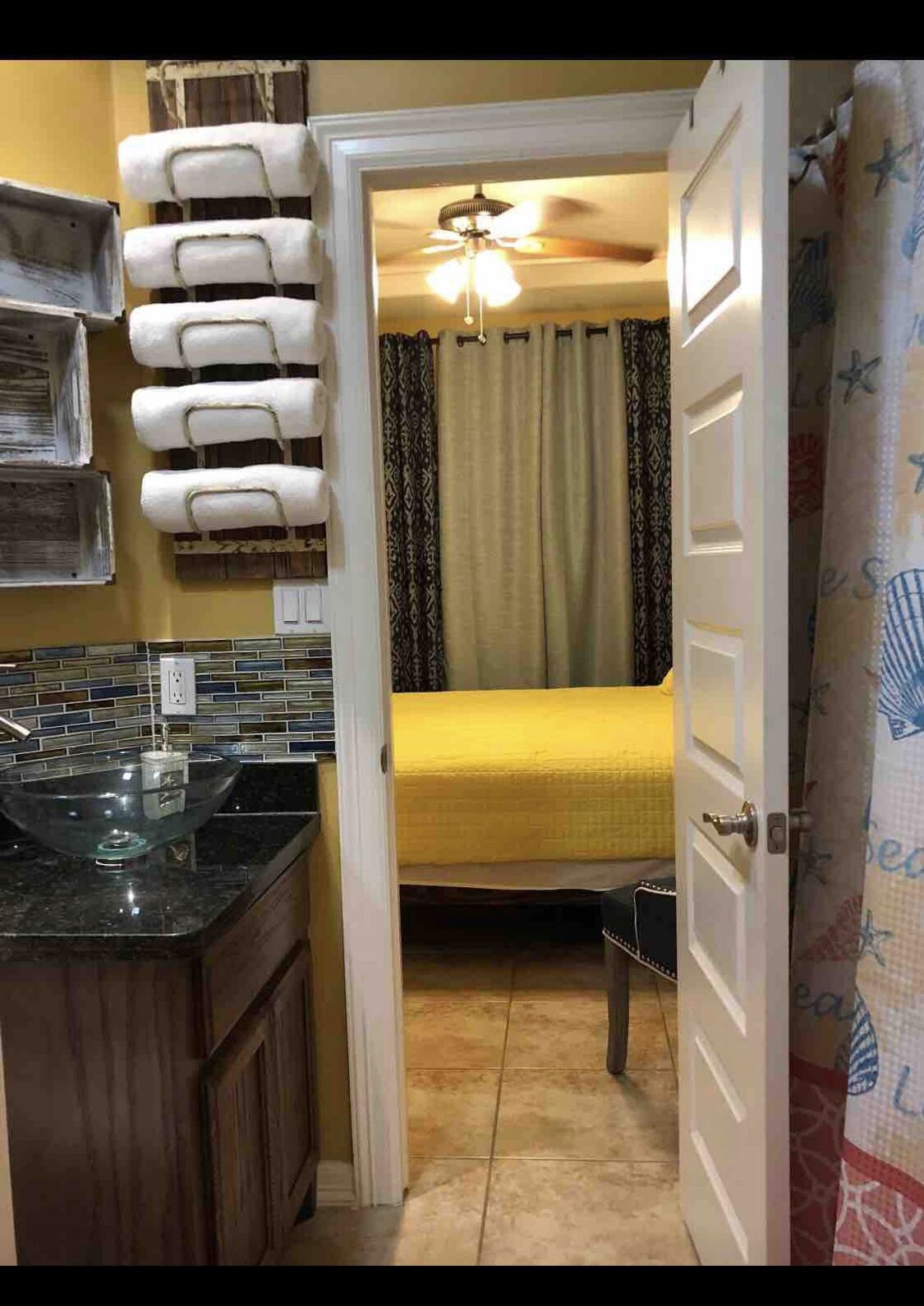 A compact kitchen with a glass bowl sink, a tiled backsplash, and a wooden cabinet. A bed with a yellow blanket and patterned curtains is visible in the adjacent room.