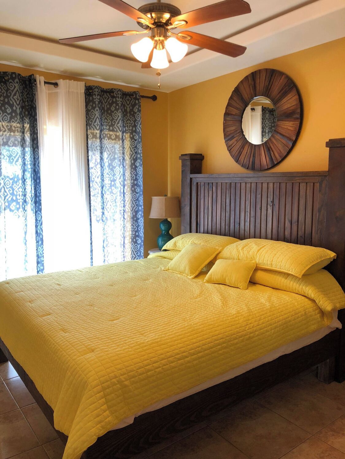 A bedroom with a bed, a ceiling fan, a round mirror, and a nightstand with a lamp.