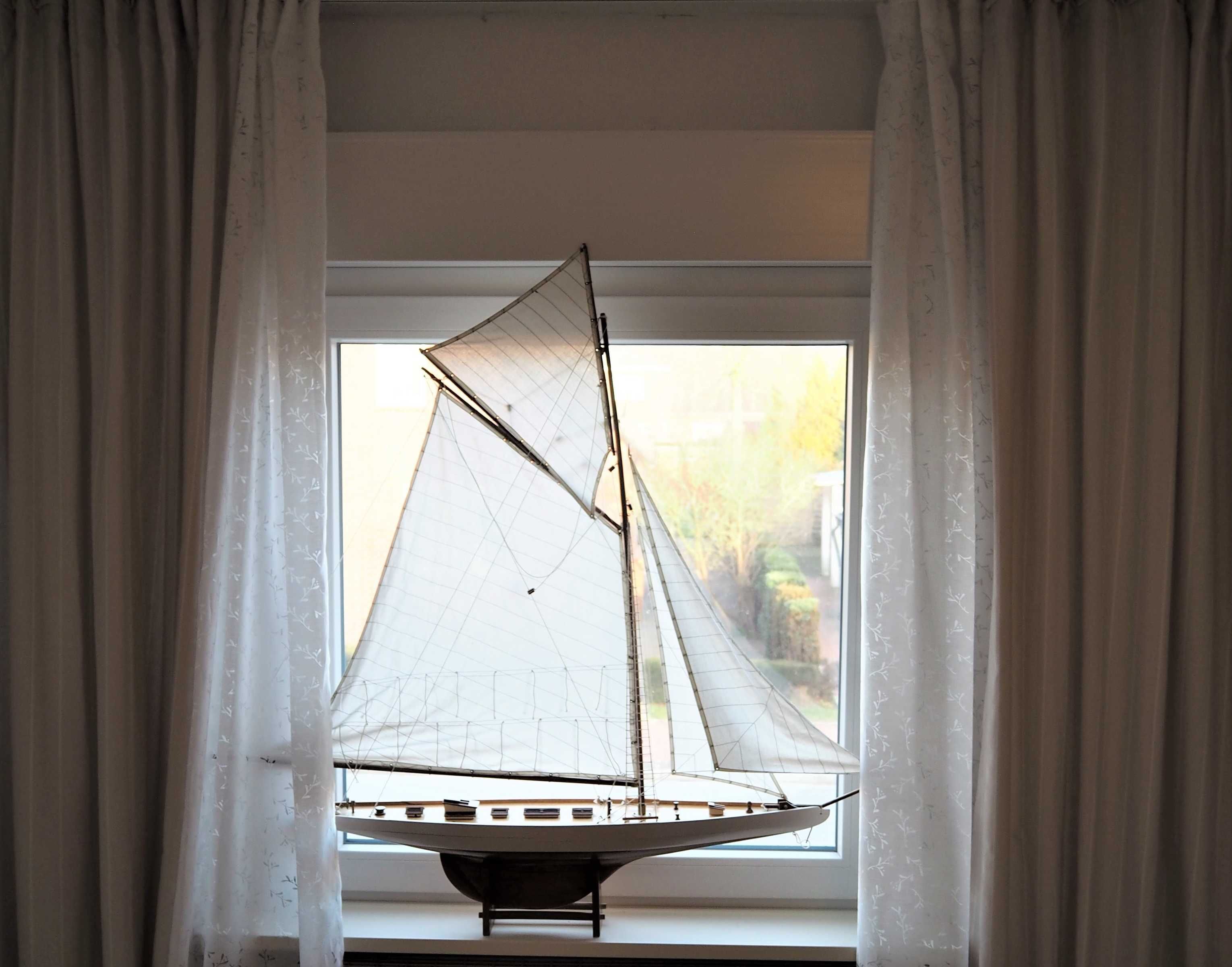 Ein Segelbootmodell ist in einem Schaufenster mit transparenten Vorhängen ausgestellt.