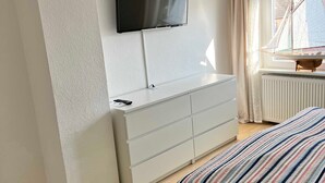 Ein modernes Schlafzimmer mit Bett, Wand-Fernseher, Kommode und einem Vorhangfenster.