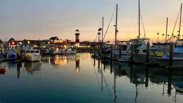 Ein Hafen im Abenddämmerung mit vertäuten Booten, einem Leuchtturm und Gebäuden im Hintergrund.