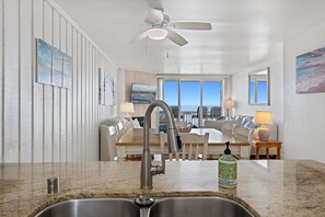 A modern kitchen with a double sink, a ceiling fan, and a view of the ocean through large windows.