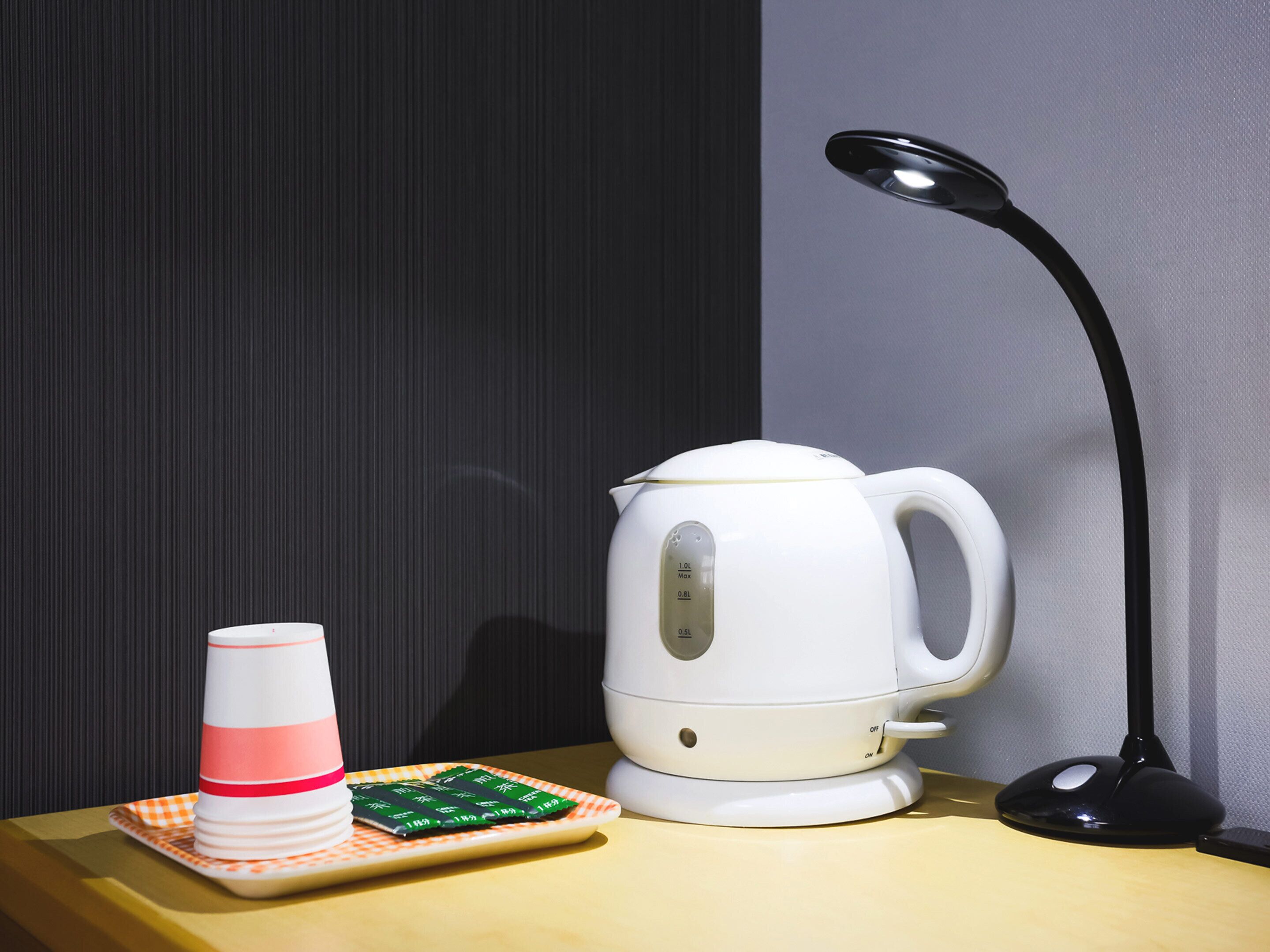 A white electric kettle, a black desk lamp, and a plate with green packets on a yellow table against a dark background.