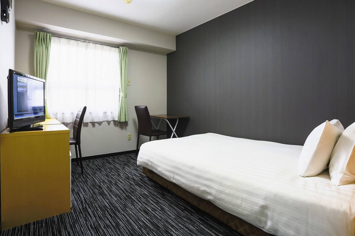 A hotel room with a bed, a desk with a computer, a chair, and a window with curtains.