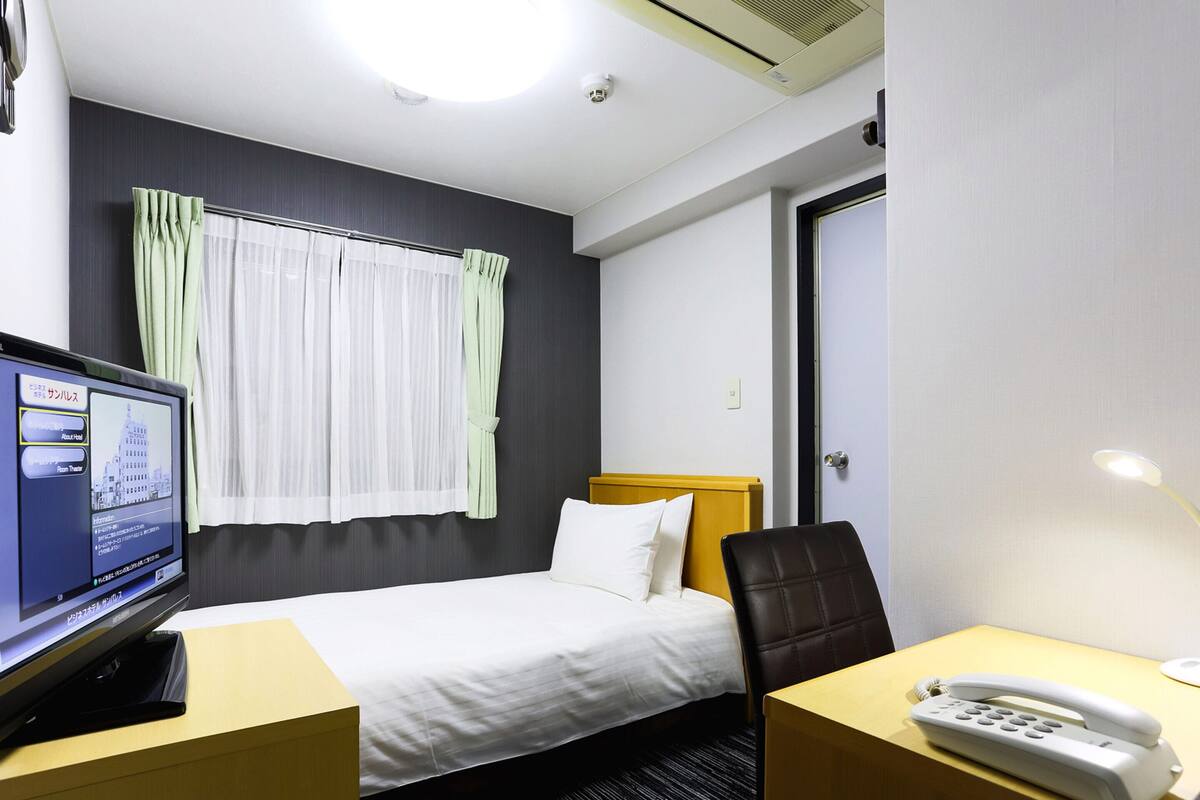 A hotel room with a bed, a desk with a phone, a TV on a stand, and a window with curtains.