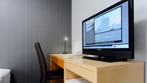 A hotel room with a bed, a desk with a computer monitor, a chair, and a small refrigerator.