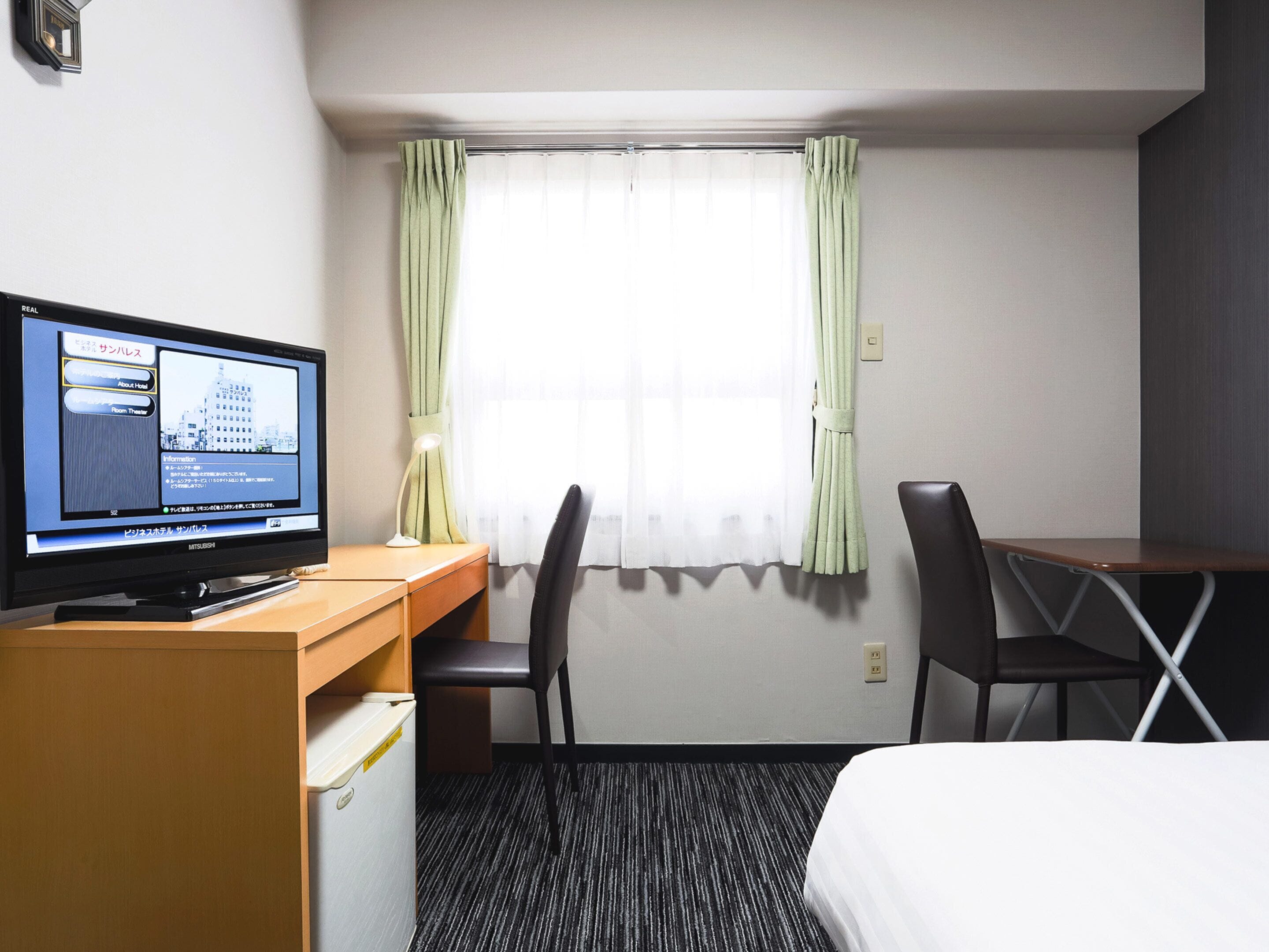 Une chambre d’hôtel comprenant un lit, un bureau avec un ordinateur, deux chaises, une fenêtre avec des rideaux et une horloge murale.