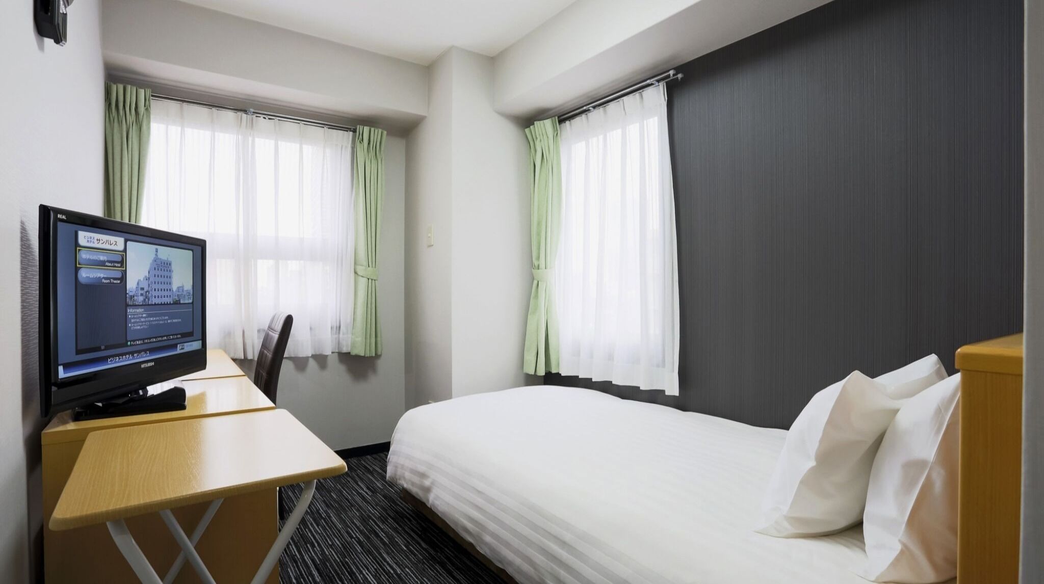 A hotel room with a bed, a desk with a computer, and two windows with curtains.