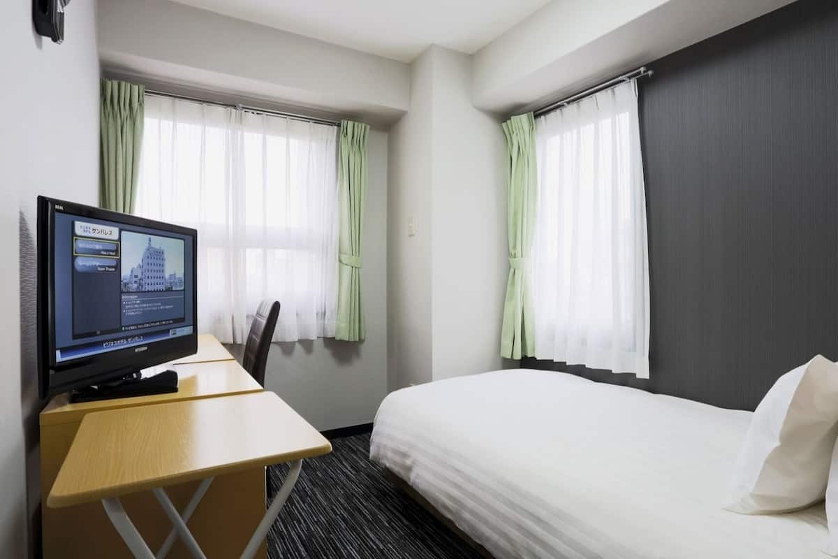 A hotel room with a bed, a desk with a computer, and two windows with curtains.