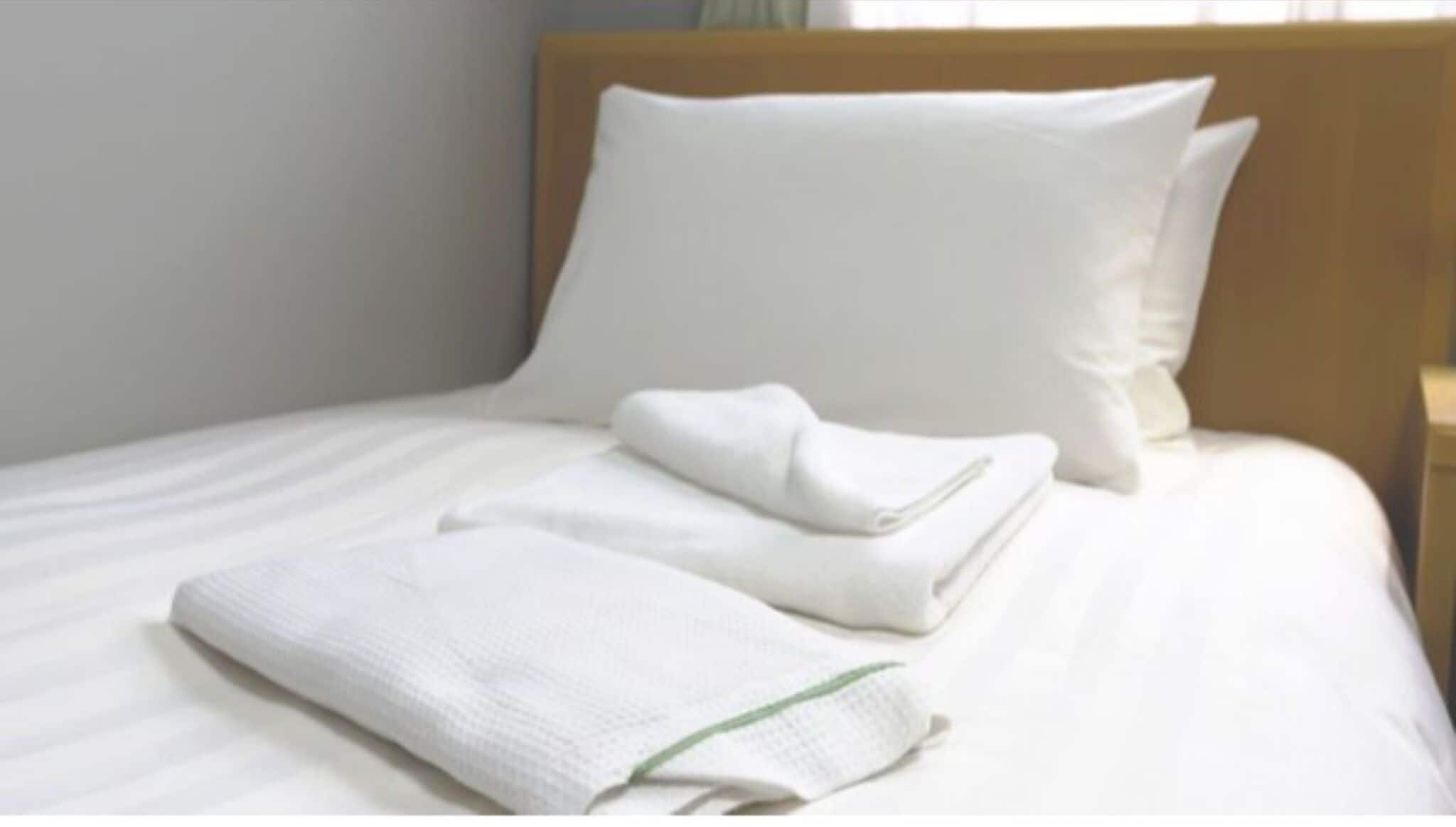 A neatly made bed with white linens and pillows, a folded towel, and a wooden headboard.
