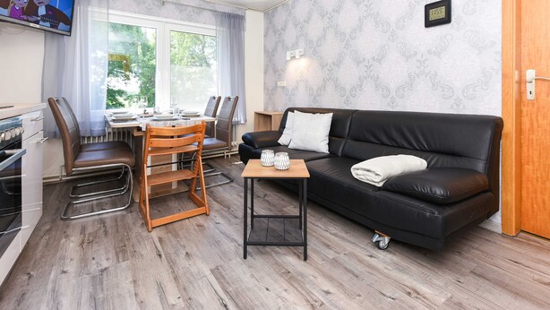 Een moderne woonkamer met een zwarte leren bank, een houten eettafel en stoelen. Tussen de bank en de eethoek staat een klein houten bijzettafeltje.