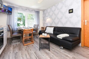 A modern living room with a black leather sofa, wooden dining table, and chairs. A small wooden side table is placed between the sofa and dining area. - Apartment "South" - Ferienhof "Tannen" (Stedesdorf)