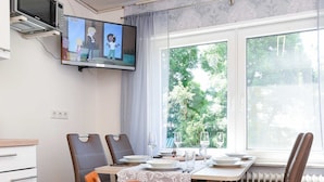 Een eetgedeelte met een tafel gedekt voor twee personen, een aan de muur bevestigde televisie en een raam met uitzicht op groen.