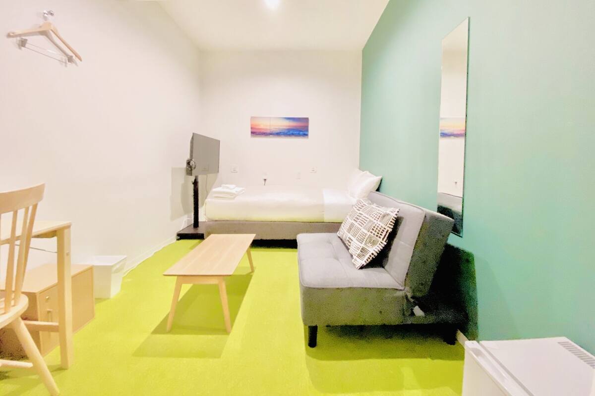 A room with a bed, a sofa, a desk, a chair, a table, a lamp, and a painting on the wall.