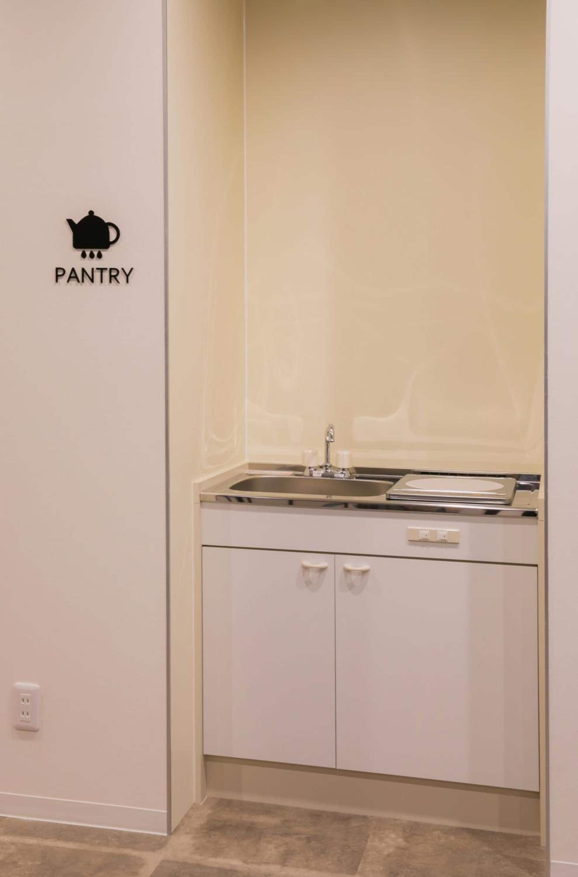 A small kitchenette with a sink, a stove, and a cabinet. There is a wall sign with a teapot icon and the word 'PANTRY' on it.