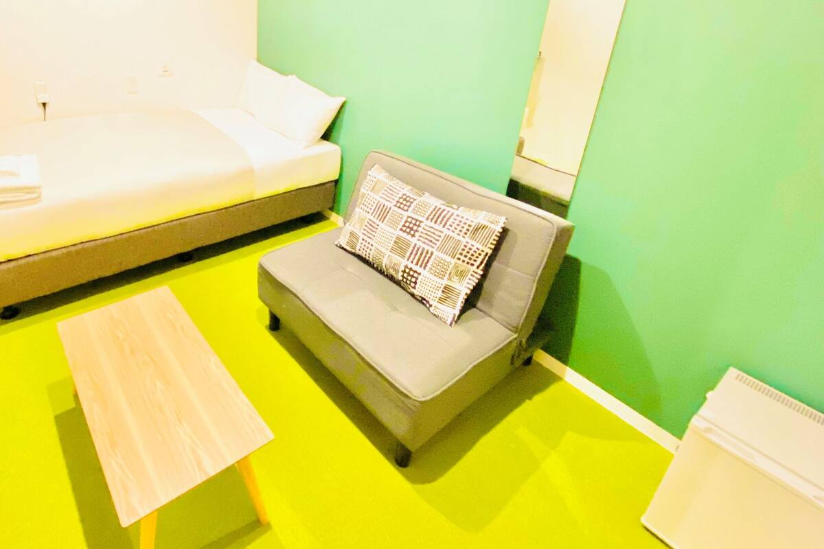 A small, brightly colored room with a green wall, a grey sofa with a patterned pillow, a white bedside table, and a painting on the wall.