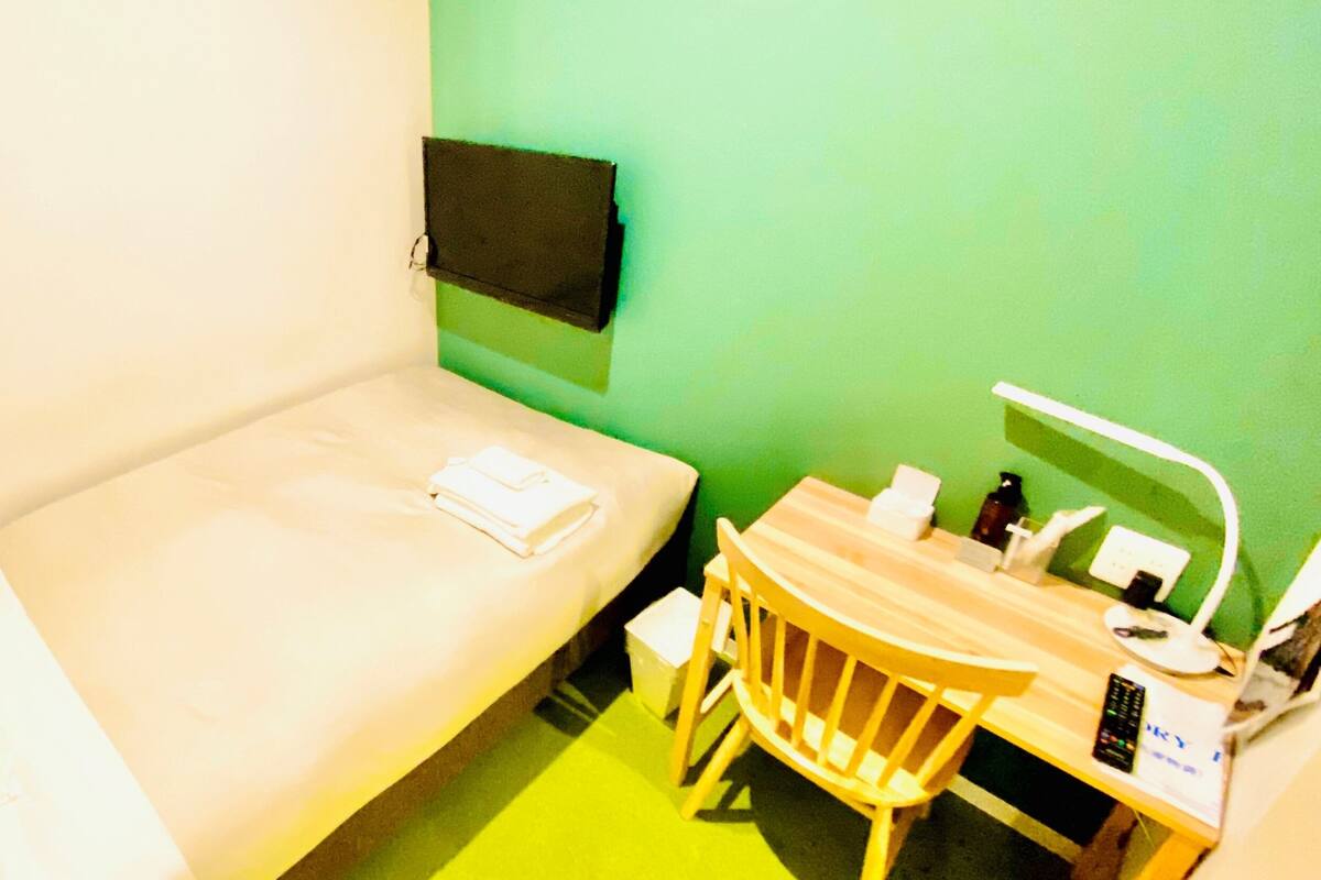 A small, single-bed room with a green wall, a wooden desk with a chair, a small table, and a TV mounted on the wall.