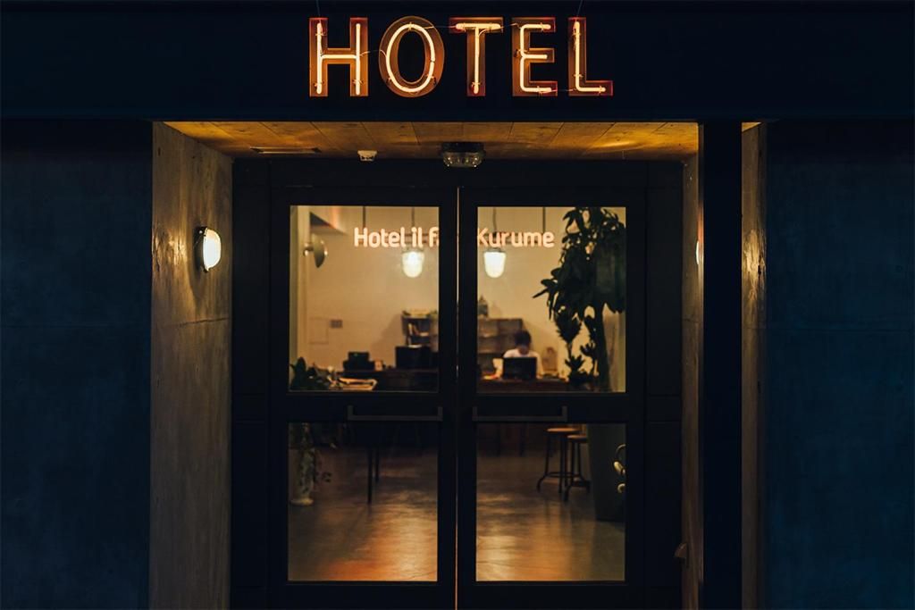 A hotel entrance with a sign reading 'HOTEL' above the doors. Inside, there are tables and chairs, and a person is visible working at a desk.