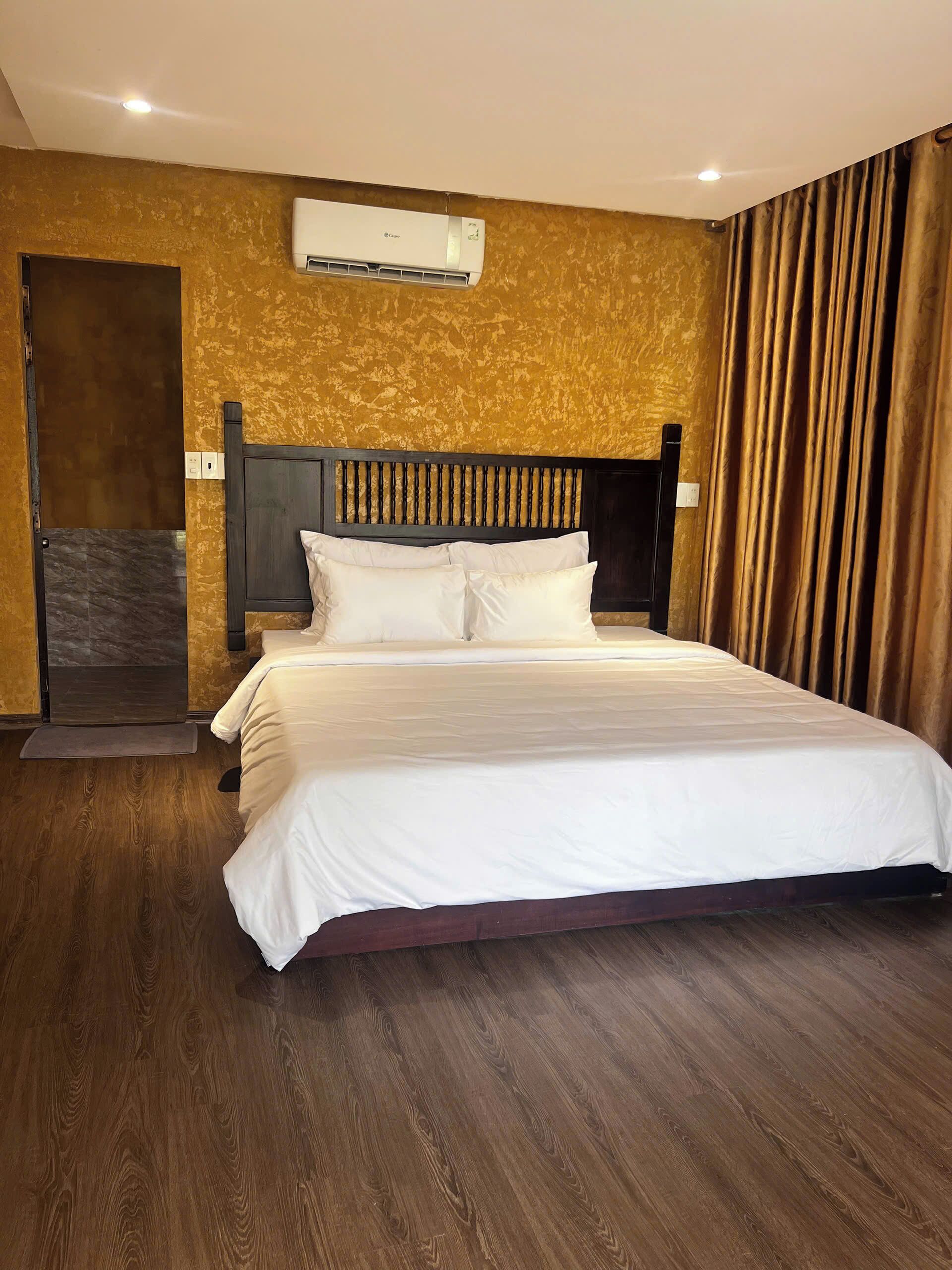 A hotel room with a wooden floor, a bed with white bedding, a wooden headboard, and an air conditioning unit on the wall.