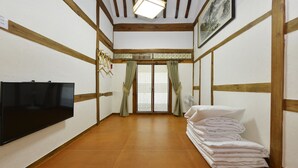 Une pièce dotée d’un plancher en bois, d’un téléviseur à écran plat fixé au mur, d’une pile de linge de lit blanc et d’une fenêtre de style traditionnel agrémentée de rideaux verts.