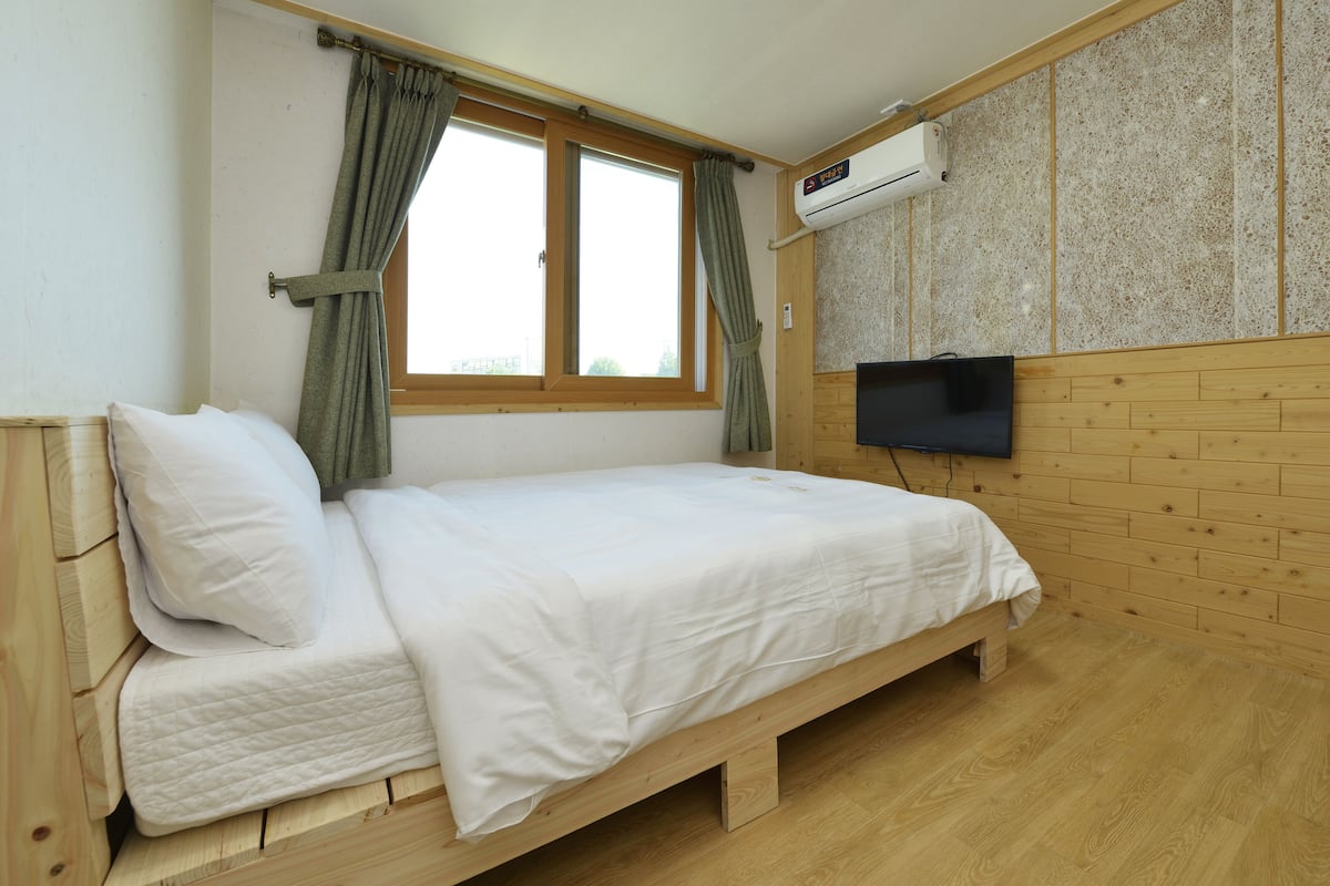 A single bed with a wooden frame, a flat-screen TV mounted on the wall, and an air conditioning unit above the TV.