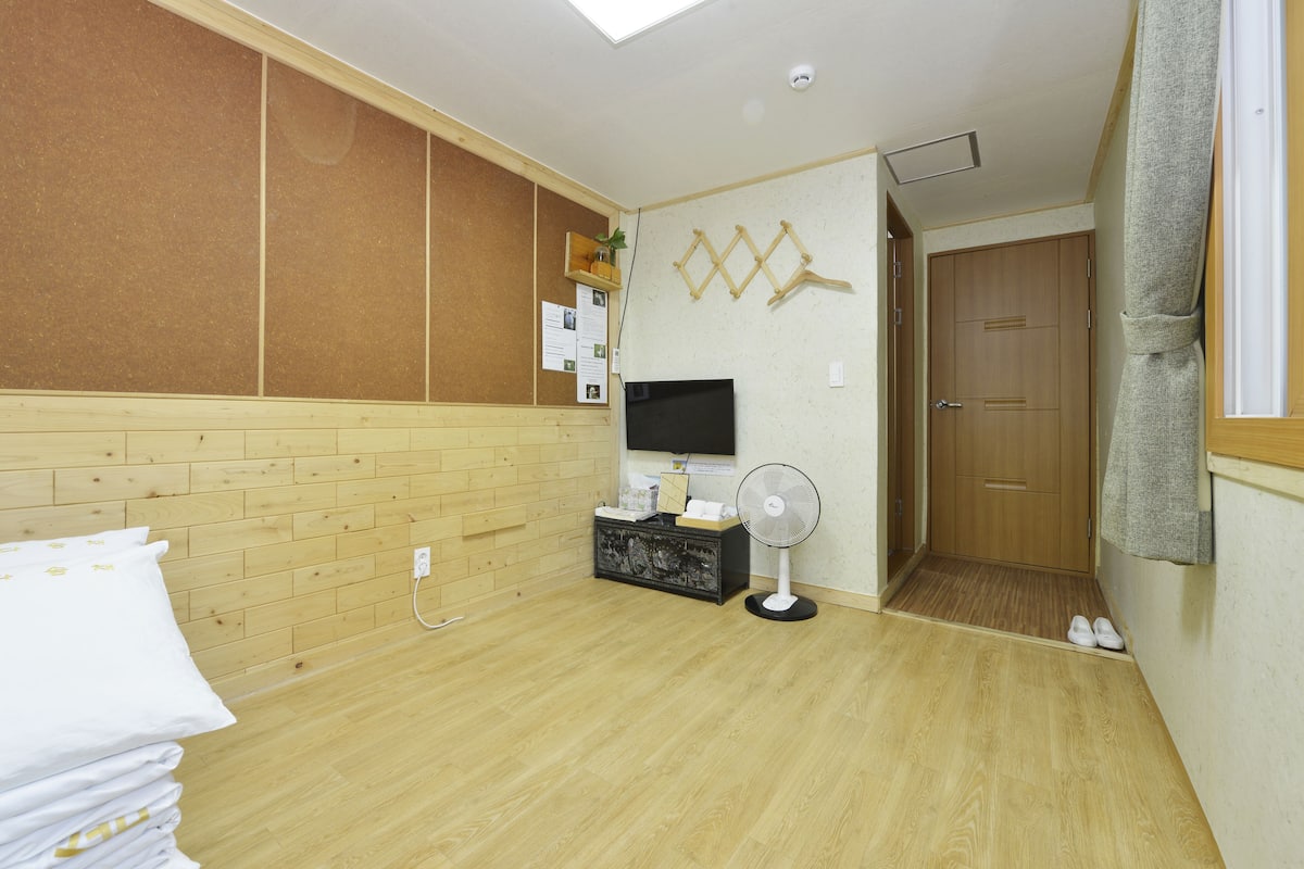 A room with a wooden floor, a TV, a fan, a corkboard with notes, and a door.