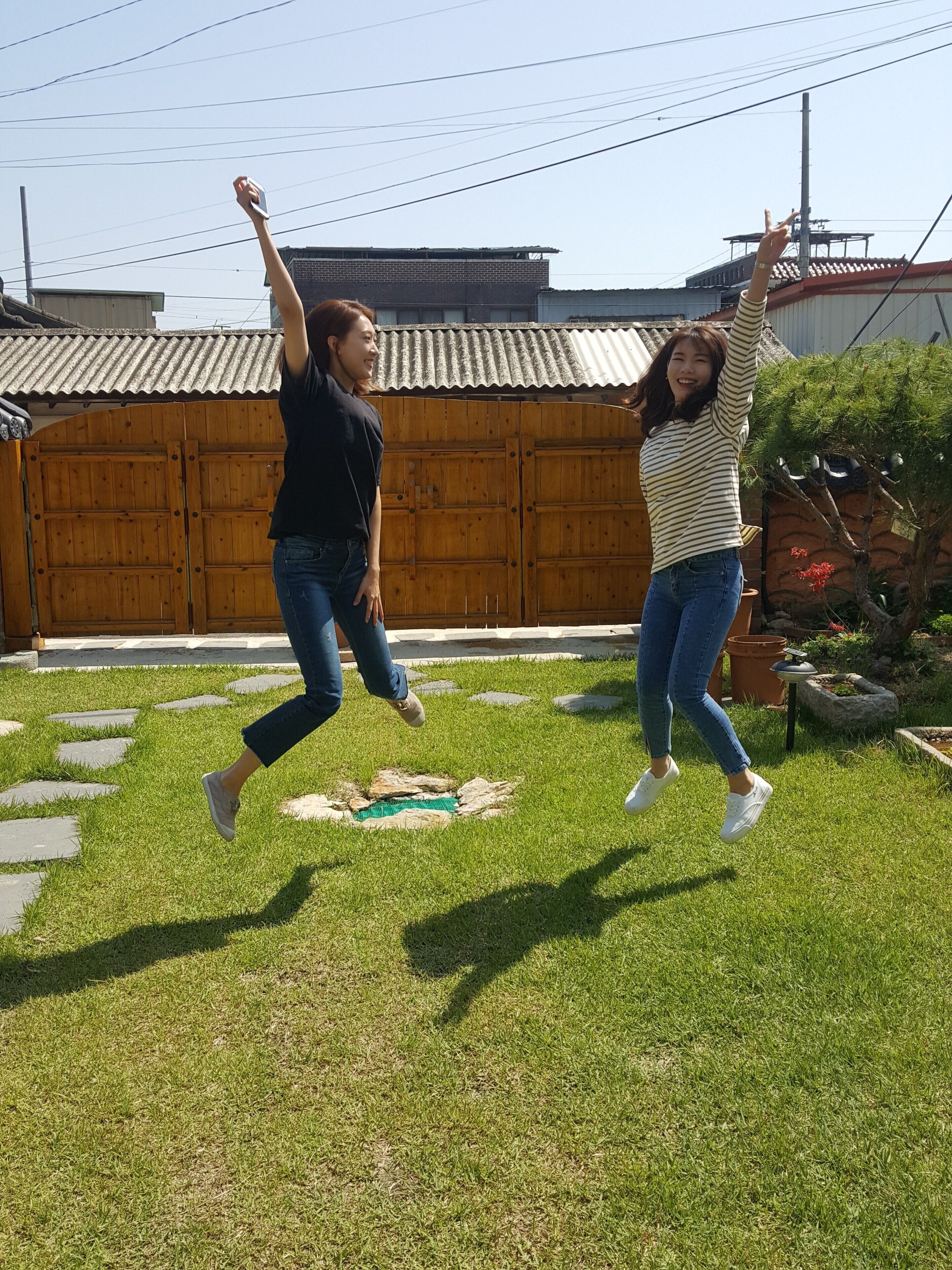 Two people jumping in a grassy yard with a wooden fence and a building in the background.