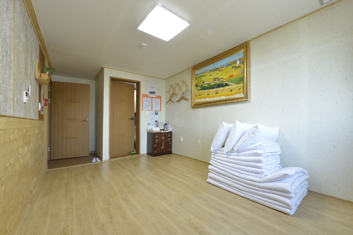 A hotel room with wooden flooring, a painting on the wall, a stack of white pillows, and a wooden cabinet.