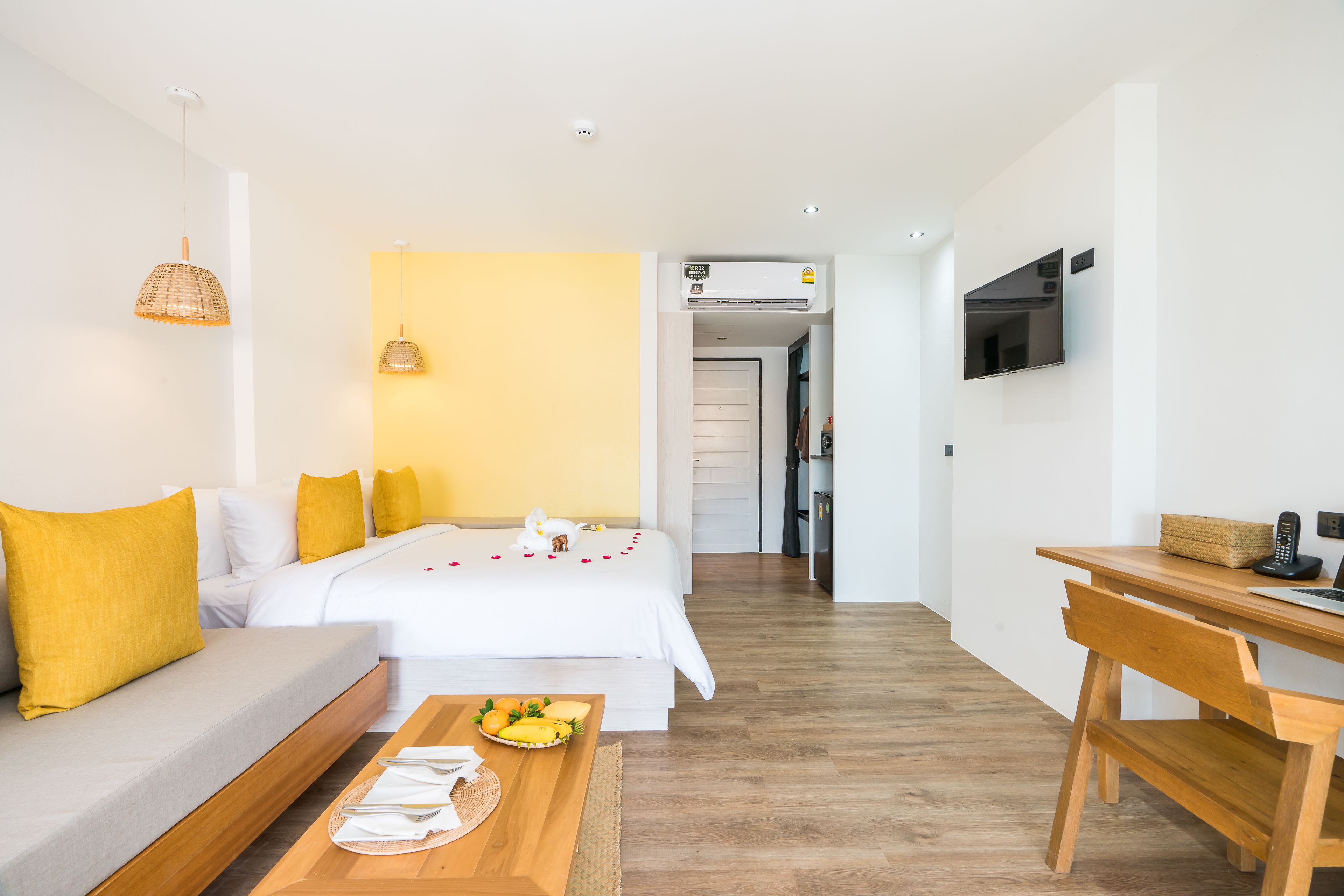 Ein modernes Hotelzimmer mit einer Couch, einem Holztisch mit Früchten, einem Bett mit weißer Bettwäsche und einem an der Wand montierten Fernseher.