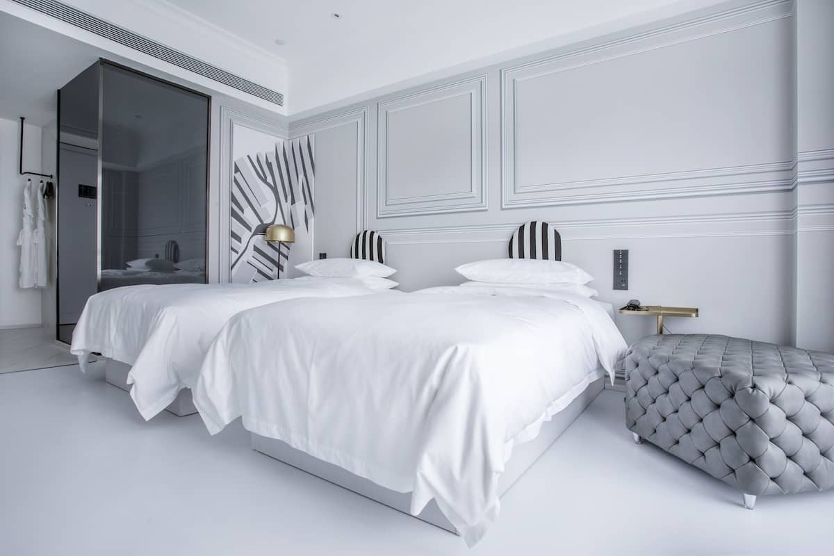 Un dormitorio moderno con dos camas, un espejo grande y un otomano acolchado.