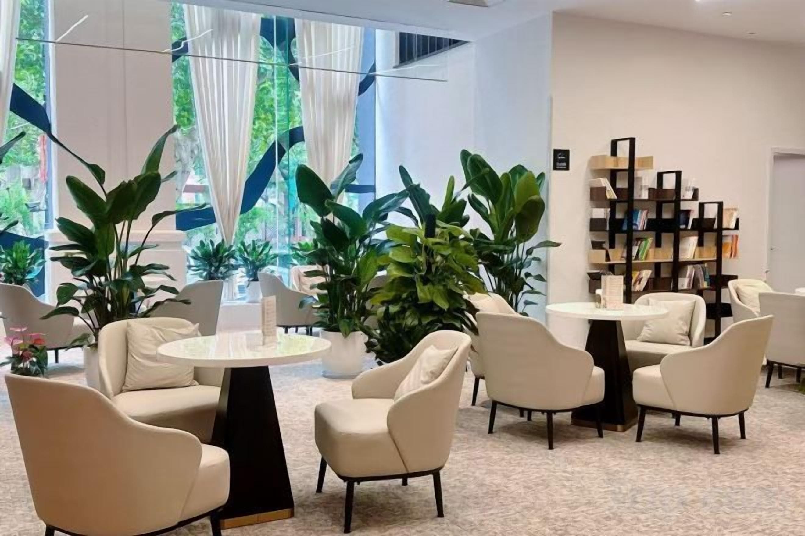 A modern lounge area with round tables and chairs, a bookshelf, and large windows with curtains.