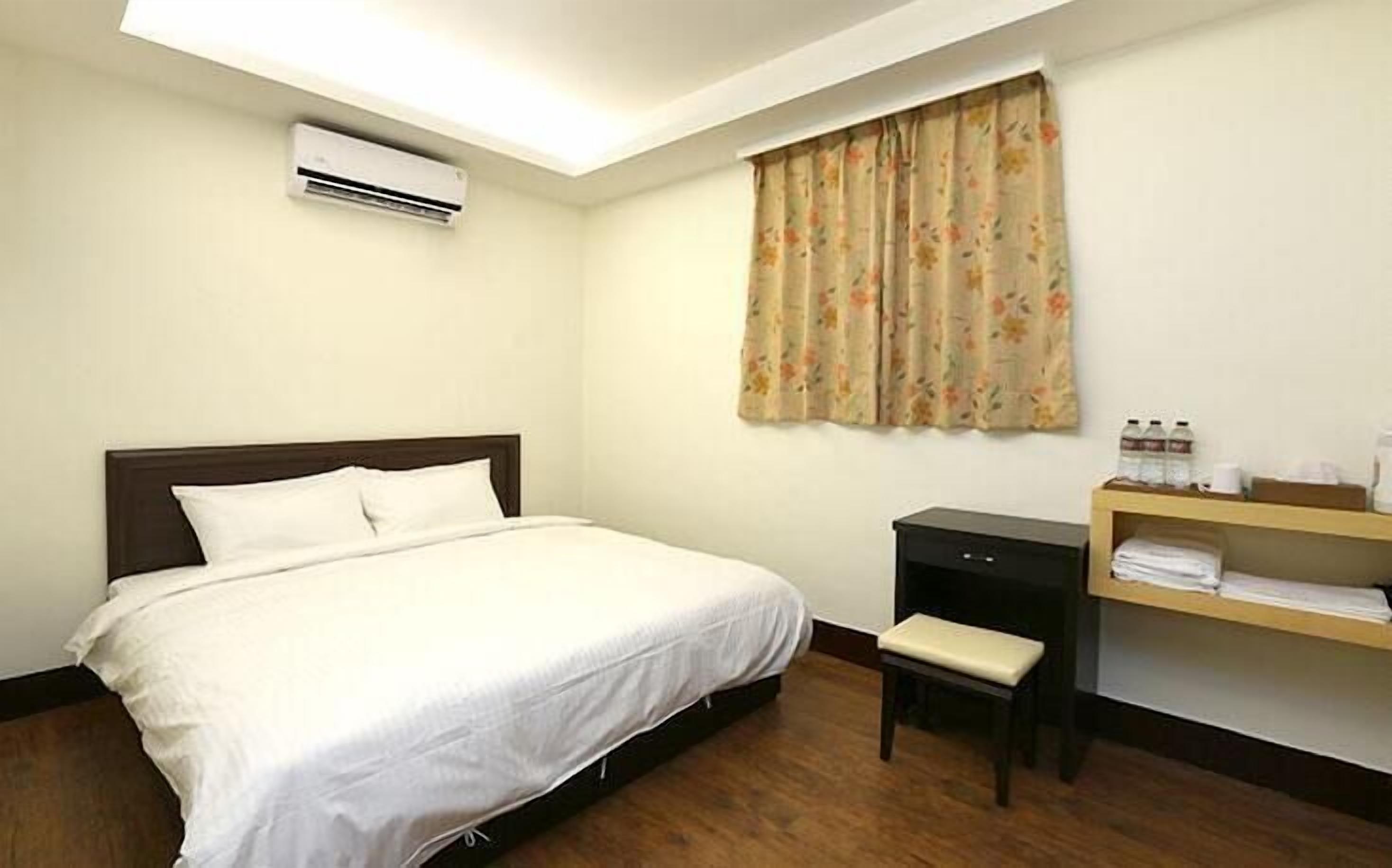 A hotel room with a bed, a small desk, a chair, an air conditioning unit, and a window with curtains.