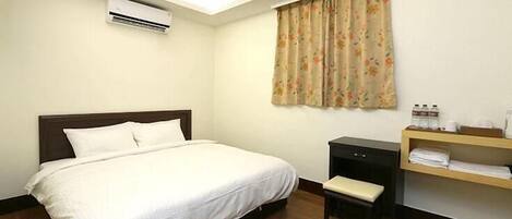 A hotel room with a bed, a small desk, a chair, an air conditioning unit, and a window with curtains.