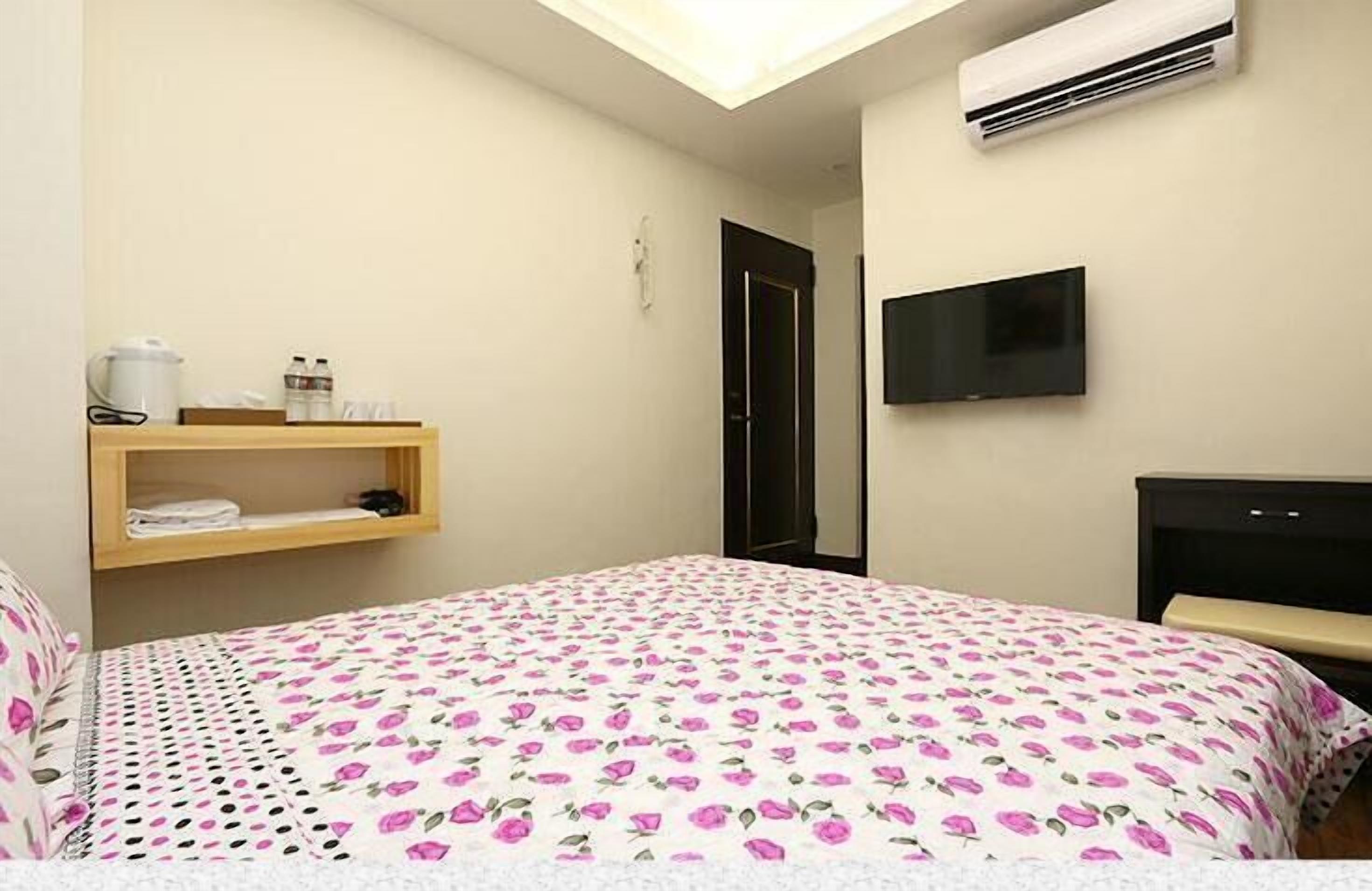 A bedroom with a bed, a TV, an air conditioner, and a shelf with toiletries.