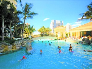 A swimming pool area with palm trees, a bridge, and people swimming.