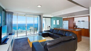 A modern living room with a large black leather sofa, a glass coffee table, and a view of the ocean through a sliding glass door. - The Towers Chevron Renaissance Holidays Gold Coast (Surfers Paradise)
