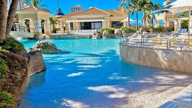 A resort pool area with clear blue water, palm trees, and lounge chairs. - The Towers Chevron Renaissance Holidays Gold Coast (Surfers Paradise)