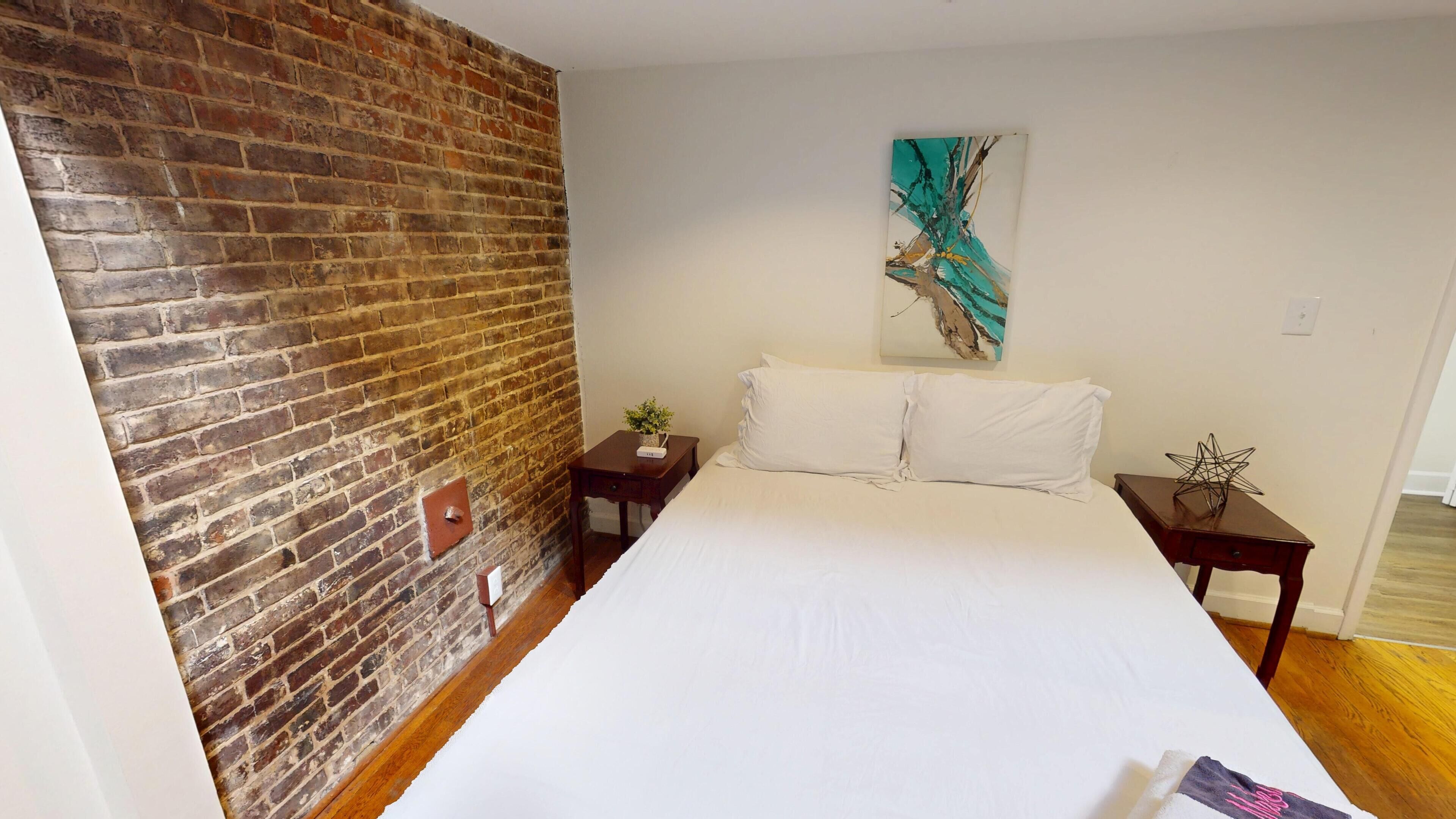 A hotel room with a brick wall, a large bed, two nightstands, and a painting on the wall.